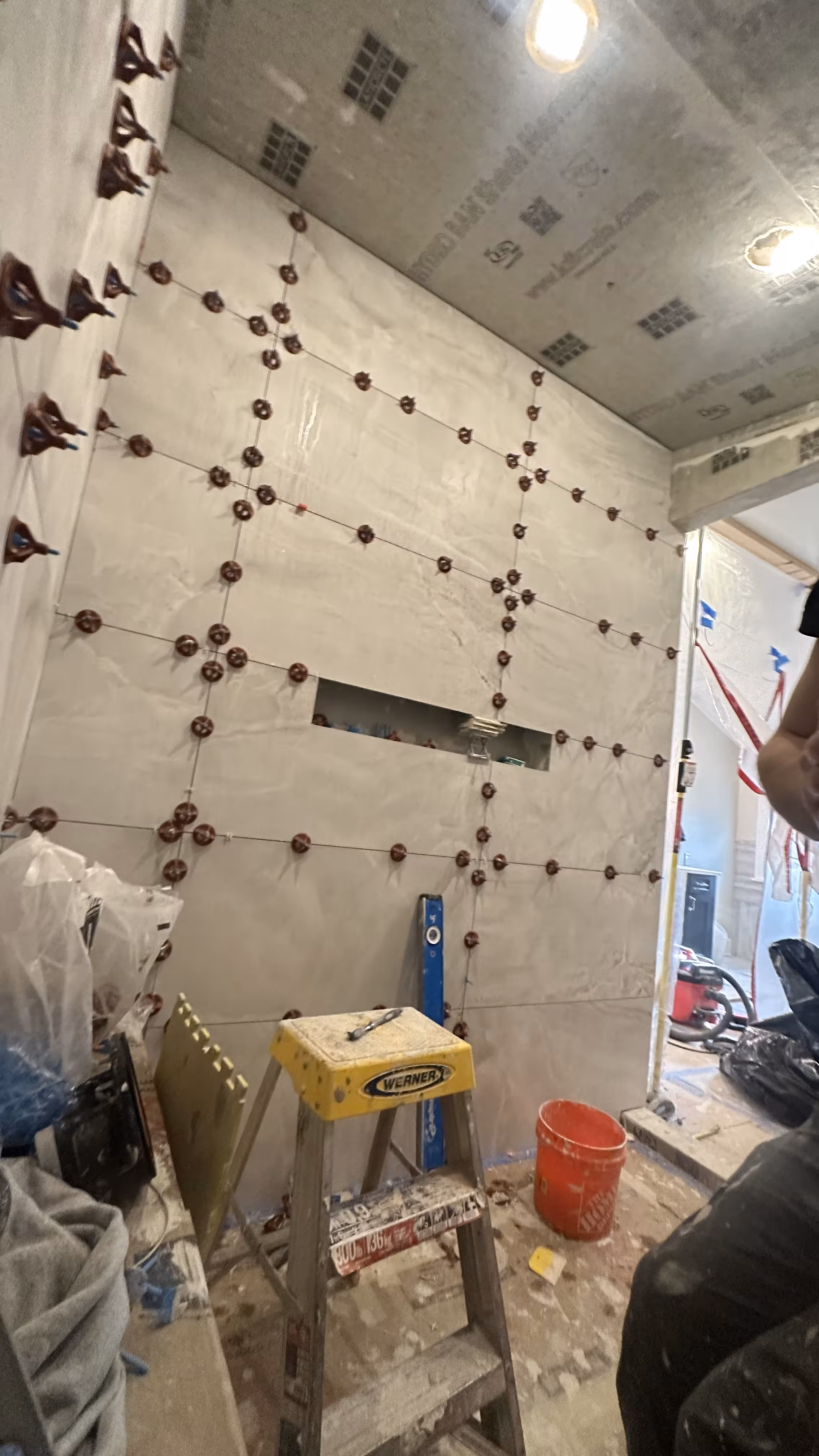 Steam shower wall mid-installation with a grid of tile leveling clips, a ladder, and a mortar bucket in Denver, CO