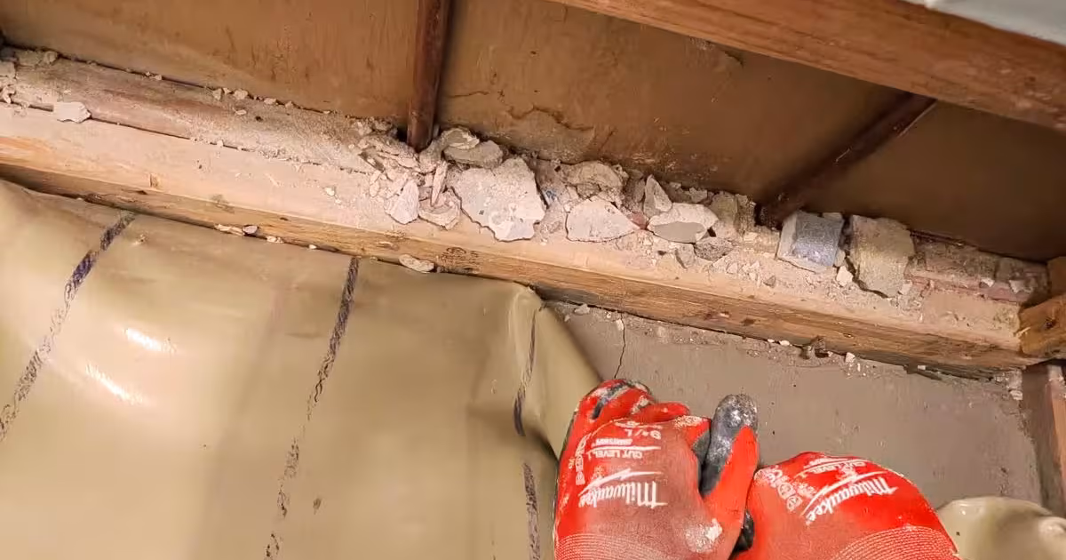 Floor joist and subfloor exposed under a shower pan in Denver, with the old PVC liner peeled back during a pan replacement