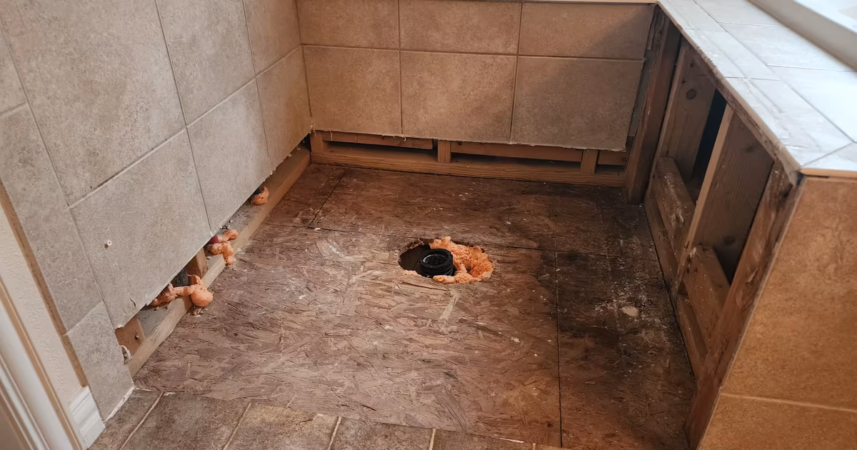 Exposed plywood subfloor and drain pipe fitting after removing damaged shower tile during a water leak repair in Aurora, CO