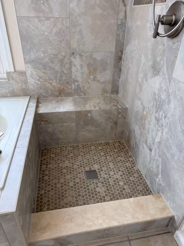 Finished walk-in shower with tiled bench, penny-round floor, and marble-look walls viewed from the tub edge in this Highlands Ranch master bath