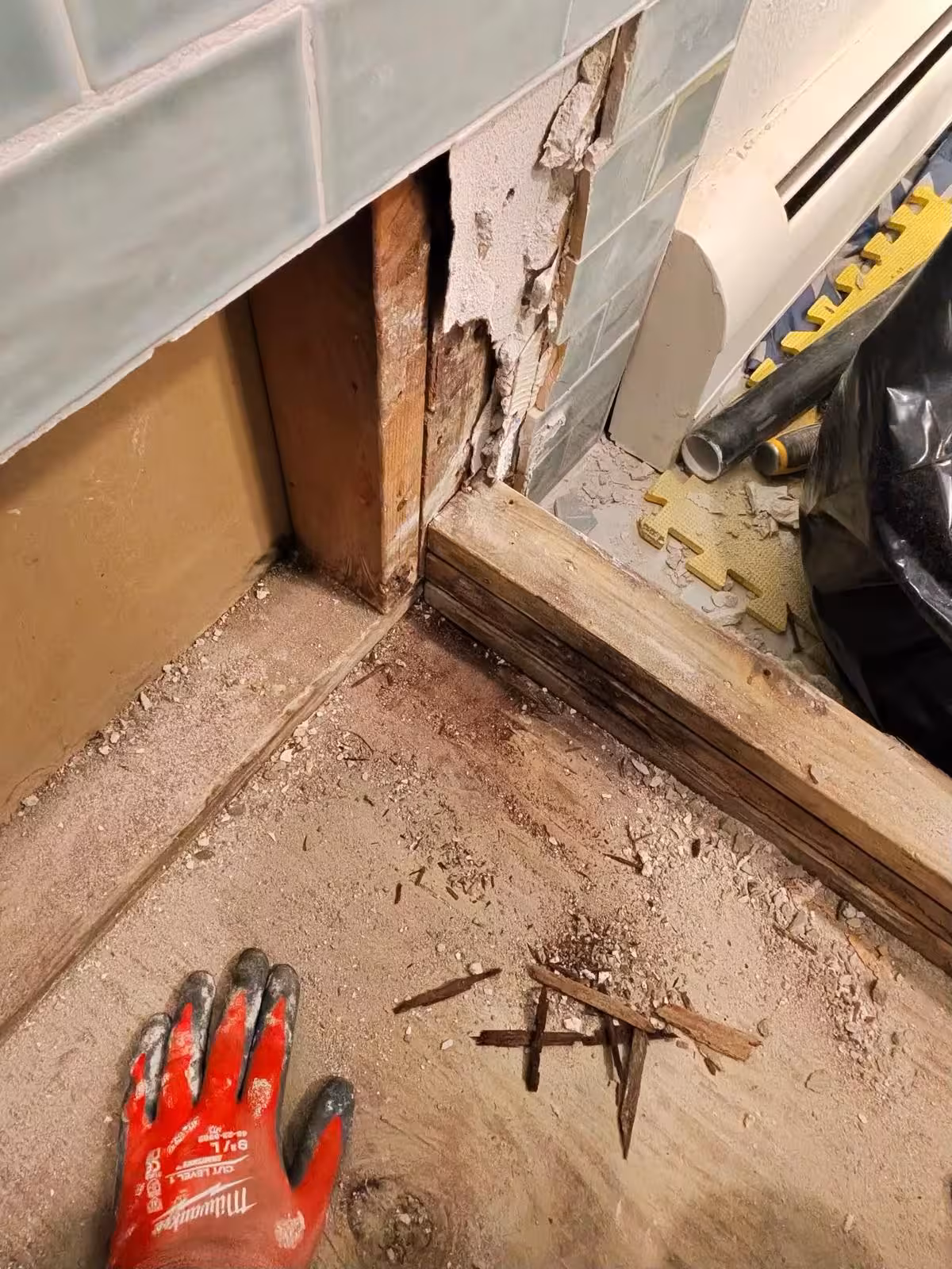 Bare subfloor and curb framing exposed at the entry of a Denver shower after the floor tile and mortar bed were removed, with the wall tile still in place above