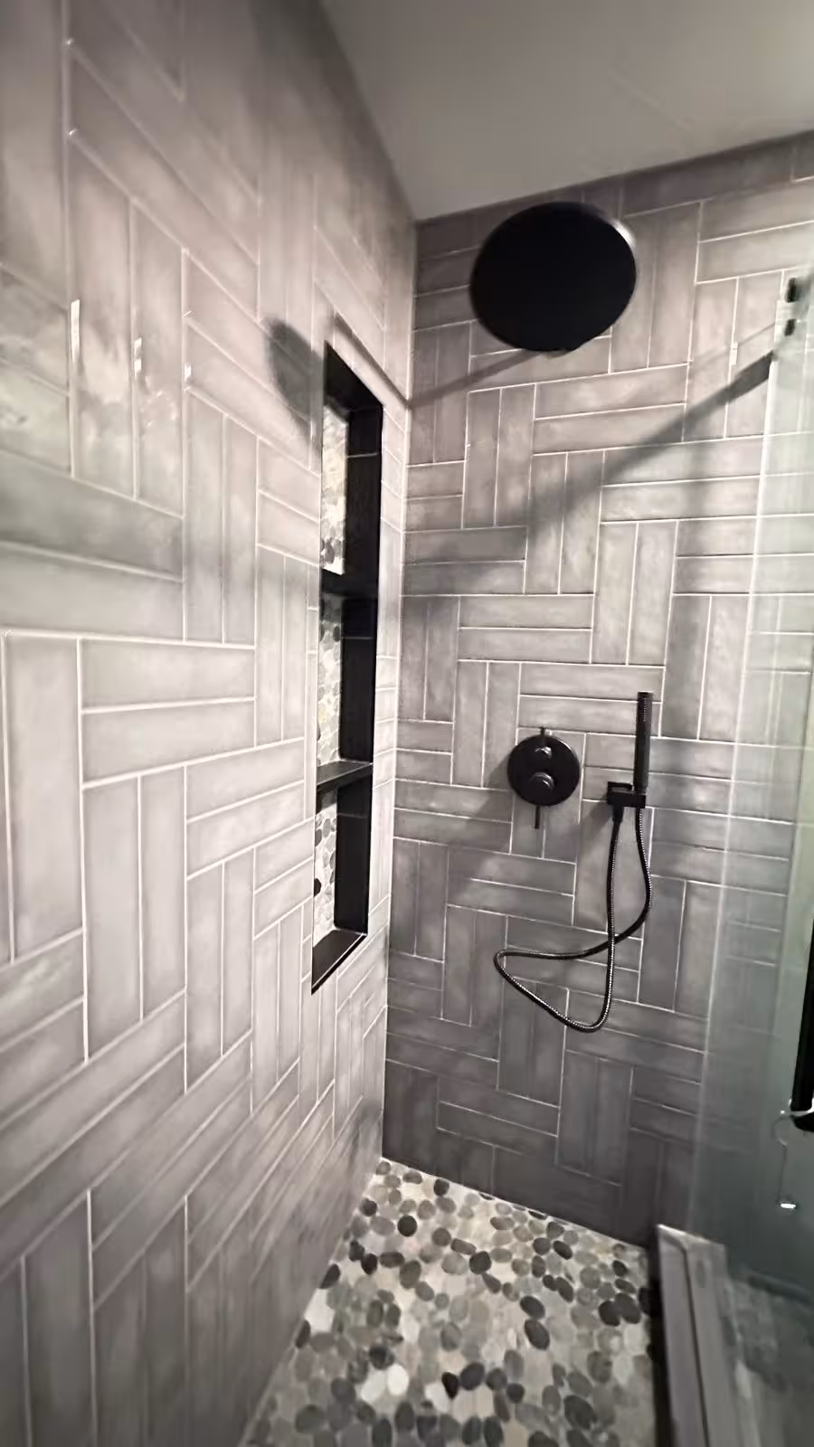 Walk-in shower corner view showing black fixtures gray herringbone tile and glass enclosure